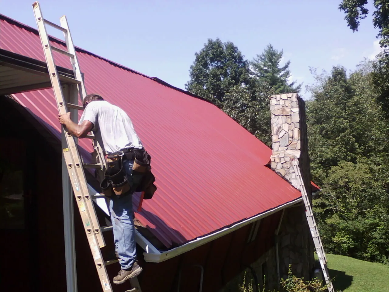 Roofer on a ladder beside a red metal roof and stone chimney — Soffit & Fascia Repair by Roofing Brothers in Lansdale